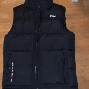 Svart dunväst från WOXO 720° - Svart dunväst från WOXO 720° med hög krage och två dragkedjefickor framtill. Västen har quiltad design och stängs med dragkedja. Perfekt för lager-på-lager och funkar till både chill och uteaktiviteter.