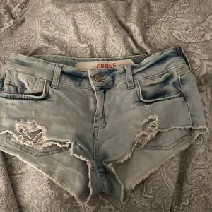Ljusblå slitna jeansshorts från Cross - Snygga ljusblå jeansshorts från Cross med råa kanter och slitna detaljer framtill. Klassisk femficksmodell med låg midja och dragkedja. Perfekta för sommaren och ger en cool, avslappnad vibe. Storlek xs men är stretchiga så passar även S