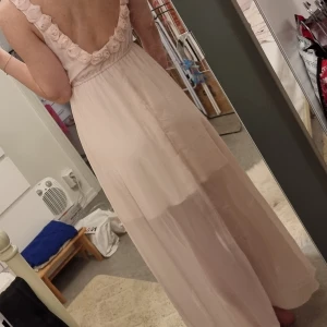 Ljusrosa långklänning Gina Tricot - Supersöt ljusrosa långklänning från Gina Tricot med volangdetaljer längs axelbanden och djup rygg. Klänningen har asymmetrisk nederkant och är tillverkad i ett tunt, flowy material som ger en romantisk vibe.