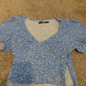 Blommig blå blus från BikBok XS - Supersöt blommig blus från BikBok i blått med små vita blommor. Modellen har v-ringning, korta puffärmar och lätt figursydd passform. Perfekt för dig som gillar romantisk och trendig stil.