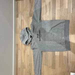 Grå hoodie Peak Performance 160 - Säljer en grå hoodie från Peak Performance med svart broderad logga på bröstet. Tröjan har huva, lång ärm och en stor magficka framtill. Perfekt för chill dagar eller när du vill ha en clean och sportig look.