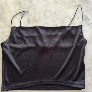 Svart satinlik bandeau topp H&M - Svart glansig bandeau topp från H&M med tunna axelband. Snygg minimalistisk look med slät yta och croppad passform. Perfekt för att styla med jeans eller kjol för en trendig vibe. Aldrig använd, lappen är fortfarande på