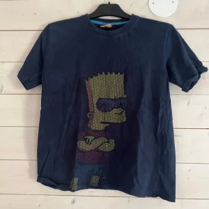 Mörkblå Bart Simpson t-shirt - Snygg mörkblå t-shirt från The Simpsons med stort tryck av Bart Simpson på framsidan. Klassisk rund hals och korta ärmar. Perfekt för dig som gillar coola och ikoniska motiv. Skön och mjuk bomullskvalitet.
