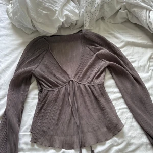 Plisserad brun blus med knyt - Supersnygg brun plisserad blus med v-ringning och långa ärmar. Blusen har ett knyt framtill under bysten och är tillverkad i ett lätt, flowy material som ger en skön känsla. Perfekt för dig som gillar en feminin och trendig look.