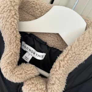 Svart quiltad dunväst från And Now This - Quiltad svart dunväst med mysigt beige foder i teddy från And Now This. Västen har hög krage, dragkedja framtill och kontrasterande detaljer i beige fuskpäls längs kanterna. Perfekt för lager-på-lager och chill höststil.