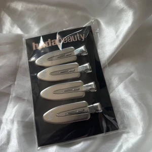 Silverfärgade hårklämmor från Huda Beauty - Fyra stilrena hårklämmor i silver från Huda Beauty. Klämmorna har en clean och modern design med Huda-loggan tryckt på ena sidan. Perfekta för att hålla håret på plats när du sminkar dig eller stylar håret.