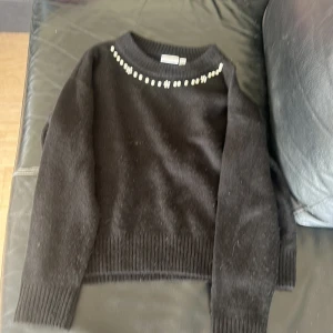 Svart stickad tröja med pärlor - Mysig svart stickad tröja med långa ärmar och pärldetaljer längs halsringningen. Perfekt för dig som vill ha en enkel men ändå lite extra look. Ribbstickade muddar vid ärmslut och nederkant ger en snygg passform.