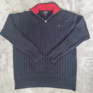 Mörkblå kabelstickad tröja Tommy Hilfiger - Kabelstickad mörkblå tröja från Tommy Hilfiger i 100% bomull. Tröjan har en halv dragkedja med röd insida på kragen och diskret logga på bröstet. Storlek XL kan passa L