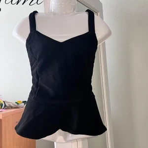 Svart peplum topp med snörning - Supersnygg svart korsettopp med breda axelband och v-ringad front. Toppen har en peplum-liknande nederdel och cool snörning i ryggen. Materialet är ribbat och ger en trendig känsla. Perfekt för dig som vill sticka ut med en edgy look.