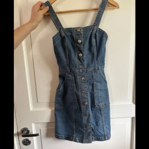 Jeansklänning från H&M Divided - Snygg jeansklänning från H&M Divided i klassisk blå denim. Kort modell med breda axelband, knappar hela vägen framtill och praktiska fickor. Perfekt för dig som gillar en avslappnad och trendig stil. Materialet är tålig jeans och passformen är normal.