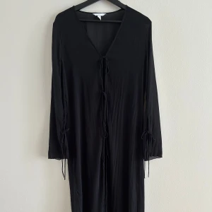 Svart långklänning med snörning - Svart långklänning i luftig polyester med v-ringning och långa ärmar. Klänningen har snygga snörningsdetaljer framtill och vid ärmsluten, vilket ger en cool och avslappnad vibe. Perfekt för dig som gillar minimalistisk stil med en twist.