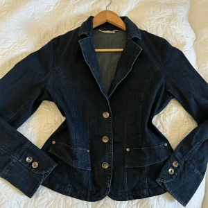 Mörkblå jeanskavaj med knappar - Snygg mörkblå jeanskavaj med markerad midja och klassisk krage. Jackan har silvriga knappar framtill, fickor med lock och dekorativa knappar vid ärmsluten. 