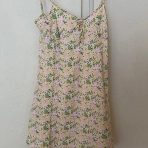 Blommig sommarklänning från H&M - Supersöt kort klänning från H&M med blommigt mönster i pastelliga färger. Tunna flätade axelband, knytning fram och öppen rygg med korsade band. Perfekt för varma dagar och ger en riktigt fräsch vibe. Har ej prislapp kvar men är oanvänd