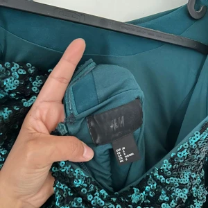 Grön paljettklänning från H&M - Snygg grön kortklänning från H&M, täckt med glittriga paljetter. Klänningen har lång ärm och en djup v-ringning i ryggen. Perfekt för dig som vill sticka ut med en festlig look. Materialet är polyester och den har dragkedja i sidan.