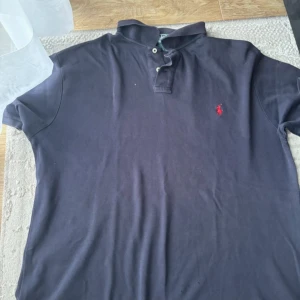 Mörkblå piké från Ralph Lauren - Klassisk mörkblå pikétröja från Ralph Lauren med röd broderad logga på bröstet. Tröjan har korta ärmar, krage och knappar framtill. Tillverkad i mjuk bomull som är skön mot huden. Perfekt för en avslappnad och stilren look. Den har jätte små hål som knappast syns vid användning.