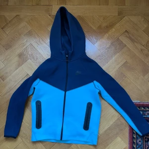 Nike tech blå med huva - Nike vindjacka i två nyanser av blått med svart dragkedja och huva. Jackan har två svarta fickor med dragkedja framtill och är tillverkad i ett mjukt syntetmaterial. Perfekt för dig som gillar sportig stil och vill ha något bekvämt och snyggt.