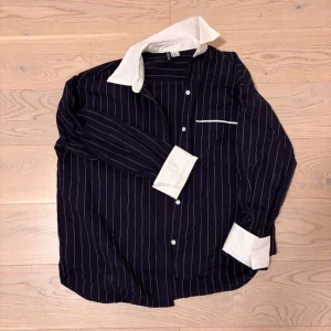 Randig skjorta med vit krage  - Snygg skjorta med vita tunna ränder från H&M. Skjortan har en kontrasterande vit krage och vita manschetter, samt knappar framtill och en bröstficka med vit kant. Perfekt för dig som gillar en klassisk men ändå trendig look.