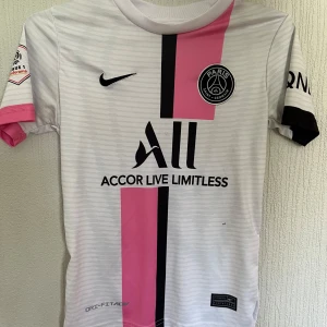 PSG Mbappé matchtröja vit/rosa Nike - Paris Saint-Germain fotbollströja med Mbappé och nummer 7 på ryggen. Vit bas med svarta och rosa vertikala ränder, svarta detaljer och klubbmärke på bröstet. Kortärmad modell i lätt och ventilerande Dri-FIT-material. Snygg och sportig design för alla PSG-fans.