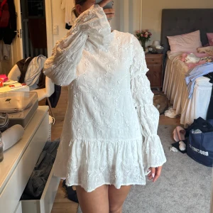 Vit kort klänning med volang - Superfin vit kort klänning med broderade blommor, volangkant nertill och vida långa ärmar. Klänningen har öppen rygg med knytband och en lös passform. 
