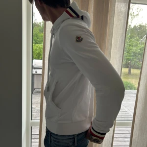 Vit Moncler hoodie med dragkedja - Snygg vit hoodie från Moncler med dragkedja, huva och snörning. Tricolore-detaljer på huva och ärmslut, samt Moncler-logga på ärmen. Tillverkad i mjuk bomull, perfekt för en clean och sportig look.