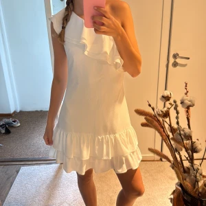 Vit one shoulder volangklänning - Supersöt vit kortklänning med one shoulder och volangdetaljer både upptill och nertill. Klänningen har en figurnära passform och är perfekt för sommarens alla tillfällen. Materialet känns lätt och luftigt, och volangerna ger en extra touch av stil.