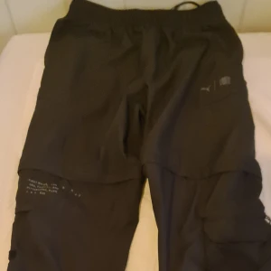 Svarta cargopants från Puma - Svarta cargopants från Puma med flera fickor och coola detaljer. Byxorna har elastisk midja och är tillverkade i ett lätt och bekvämt material, perfekt för en streetwear-look. Snygga tryckdetaljer på benen och klassisk Puma-logga på fickan.