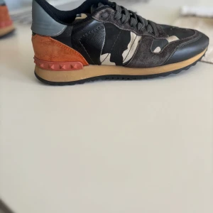 Valentino Rockrunner sneakers i camouflage  - Snygga Valentino Rockrunner sneakers med ett coolt camouflagemönster. Skorna har en kombination av svart, grått och orange mocka och skinn, samt en unik nitar-detalj på hälen. Perfekta för en trendig look. Skorna är i bra skick och är storlek 39 passar 40. OBS varken skobox eller dustbag ingår. Pris går att diskutera vid snabb affär.