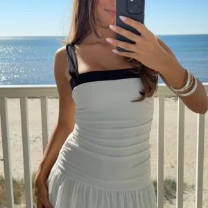 Vit och svart ärmlös sommarklänning - Supersöt vit klänning med svarta breda axelband och kant upptill. Den är ärmlös, har en tight överdel med rynkade detaljer och en lätt utställd kjol. Perfekt för varma dagar och har en fräsch, stilren look.