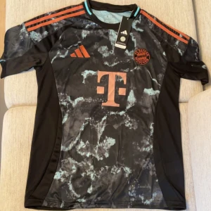 Bayern München Adidas fotbollströja - Snygg Bayern München fotbollströja från Adidas med svart och turkosmönstrad front, röda detaljer och logga. Tröjan har korta ärmar, klassiska Adidas-ränder på axlarna och är tillverkad i lätt funktionsmaterial. Perfekt för dig som vill sticka ut på planen.