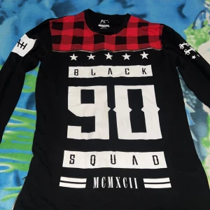 Svart långärmad tröja med röd-rutigt mönster - Cool svart långärmad tröja med stora vita tryck framtill, texten 'BLACK SQUAD 90' och romerska siffror. Röd och svart rutig detalj upptill vid axlarna. Tryck även på ärmarna. Perfekt för en streetwear-look.