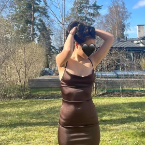 Parisian Petite satinklänning  - Elegant brun satinklänning med smala axelband och en smickrande tight passform. Klänningen har en slits baktill och en mjuk glansig yta som ger en lyxig känsla.