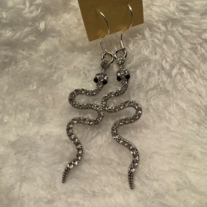 Glittriga ormformade örhängen - Säljer ett par unika örhängen i form av ormar, täckta med glittriga stenar. De är silvriga och har en böljande design som verkligen sticker ut. Perfekta för dig som vill ha något annorlunda och snyggt till din stil. Krokarna är i sterling silver 