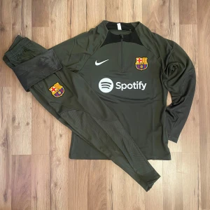 FC Barcelona träningsöverdel Nike - Mörkgrön långärmad träningsöverdel från Nike med FC Barcelona klubbmärke och Spotify-logga på bröstet. Tröjan har dragkedja vid halsen och svarta detaljer på axlarna. Perfekt för fotbollsträning eller support.