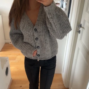 Grå stickad kofta med knappar - Mysig grå stickad kofta med stora svart/blåa knappar framtill. Koftan har en djup v-ringning och lång ärm, perfekt för lager-på-lager. 