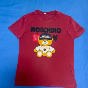 Vinröd t-shirt med nallemotiv från Moschino - Säljer en vinröd t-shirt från Moschino med ett stort broderat nallemotiv på framsidan. T-shirten har korta ärmar och är gjord i bomull. Perfekt för dig som gillar lekfulla och unika designer! Priset går och diskuteras och kan sänkas till en väldigt bra pris