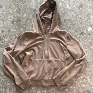 Beige hoodie i velour från Lindex - Säljer en beige hoodie i mjuk velour från Lindex. Tröjan har dragkedja, huva och en stor ficka framtill. Dekorerad med glittriga stenar i form av ett L på bröstet. Perfekt för en chill och avslappnad stil.