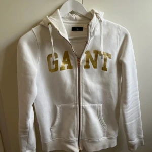 Vit hoodie med dragkedja från GANT - Vit hoodie från GANT med guldigt tryck framtill och dragkedja. Tröjan har huva med snörning och två fickor framtill. Perfekt för en avslappnad stil och enkel att matcha med det mesta.