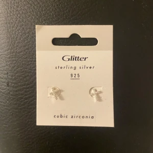 Små örhängen i silver från Glitter - Säljer ett par små kors formade örhängen i äkta sterling silver 925 från Glitter, dekorerade med cubic zirconia-stenar. Perfekta för dig som gillar diskreta och eleganta smycken i silver.