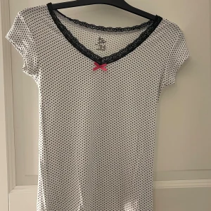 Vit prickig topp med spets och rosett - Söt vit topp med små svarta prickar, svart spetskant vid v-ringad hals och en liten rosa rosett framtill. Den är kortärmad och jätte skön i materialet 