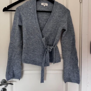 Ljusblå omlottkofta från Gina Tricot - Superfin ljusblå stickad omlottkofta från Gina Tricot i ullblandning. Koftan har långa ärmar, knytband i midjan och en mjuk, mysig känsla. Perfekt för lager på lager under kyliga dagar.