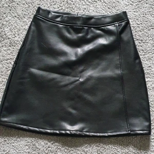 Svart skinnkjol - Säljer en svart kort skinnkjol med enkel design. Kjolen har en rak passform och är tillverkad i ett material som ser ut som skinn. Perfekt för dig som vill ha en stilren och edgy look. Den är har ett mjukare material på insidan