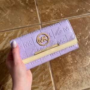 Ljuslila plånbok från Michael Kors - Säljer en snygg ljuslila plånbok från Michael Kors med präglad logotyp och gulddetaljer. Plånboken har flera kortfack, fönster för ID, myntfack med dragkedja och stängs med tryckknapp. Köpt second hand men aldrig använt själv. Det finns lite slitning inuti (bild 3&5). 