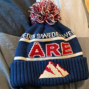 ÅRE MÖSSA (np 749kr) - Snygg blå mössa från Robin Ruth med texten 'Åre' och en färgglad tofs på toppen. Mössan har ett broderat bergsmotiv och är tillverkad i 100% akryl. Perfekt för kalla dagar!