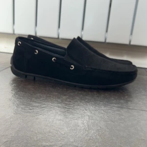 Svarta mocka loafers - Säljer ett par stilrena svarta loafers i mocka med diskreta metalldetaljer vid sidorna. Skorna har en klassisk design med rund tå och är perfekta för en clean och avslappnad look.