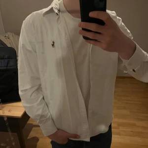 Vit skjorta från Ralph Lauren - Klassisk vit skjorta från Ralph Lauren med broderad logga på bröstet. Skjortan har lång ärm, normal passform och knappar framtill. Perfekt för en stilren och enkel look.