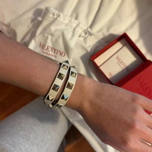 Armband med nitar från Valentino - Snyggt armband från Valentino med silverfärgade nitar. Armbandet är i en ljus beige ton och kommer i sin originalförpackning. Perfekt för att ge en edgy touch till din outfit. Kvitto och alla tillbehör finns