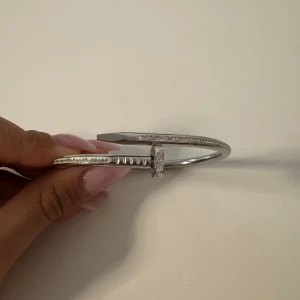 Silverfärgat armband med stenar och hjärtdetalj - Säljer ett snyggt armband i silverfärg med gnistrande stenar längs ovansidan. Armbandet har en unik design som påminner om en böjd spik och avslutas med en hjärtformad detalj. Perfekt accessoar för att lyfta din stil. Två stenar saknas mrn inget som syns🩷🩷