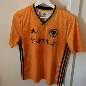 Wolves fotbollströja från Adidas - Snygg orange fotbollströja från Wolves med svart Adidas-logga och klubbmärke på bröstet. Tröjan är från 2018 och sponsortryck 'silverbug' på framsidan. Klassisk V-ringning med svart och gul kant.