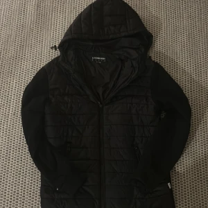 Svart jacka från Lindbergh - Snygg svart jacka från Lindbergh med huva och dragkedja. Jackan har en quiltad design och långa ärmar. Perfekt för kyligare dagar. Pris går att diskuteras vid snabb affär.