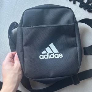 Svart axelväska från Adidas - Säljer en stilren svart axelväska från Adidas med justerbar rem och dragkedja. Perfekt för att bära med sig det nödvändigaste. Användbart i flygplatsen. Adidas-loggan i vitt pryder framsidan.